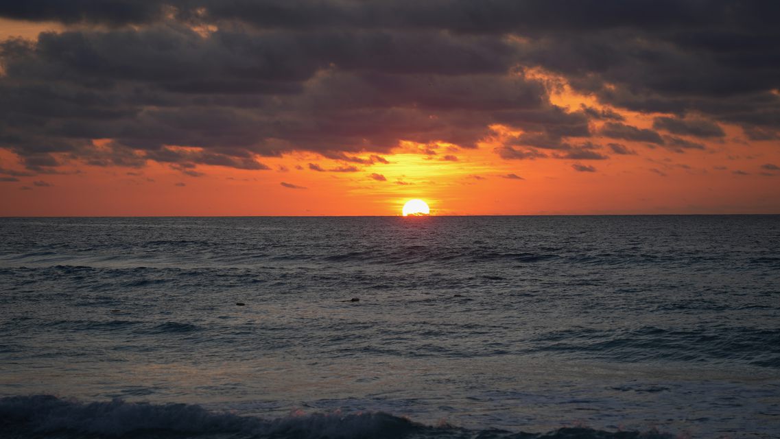 The sun low on the sea horizon under dark clouds lit orange
