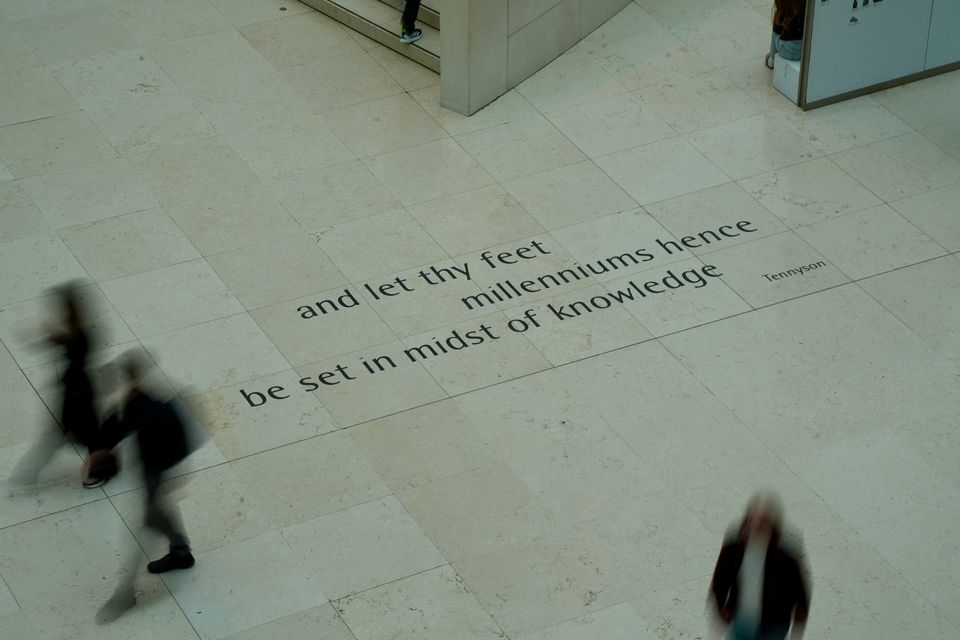 Blurred figures crossing a pale stone floor carved with a line of verse