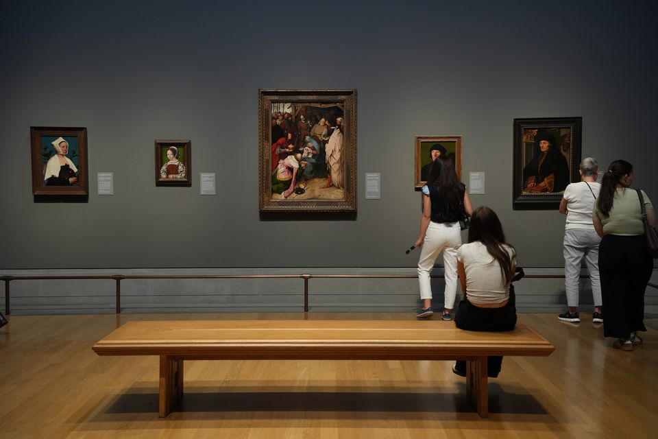 Visitors looking at framed paintings in a blue-grey gallery, a wooden bench in front