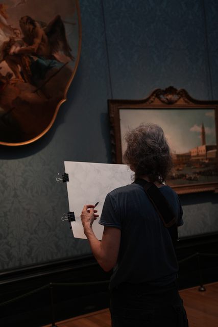A visitor sketching in front of paintings in a gallery, seen from behind