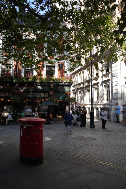 A red pillar box on a paved city square with trees and pedestrians