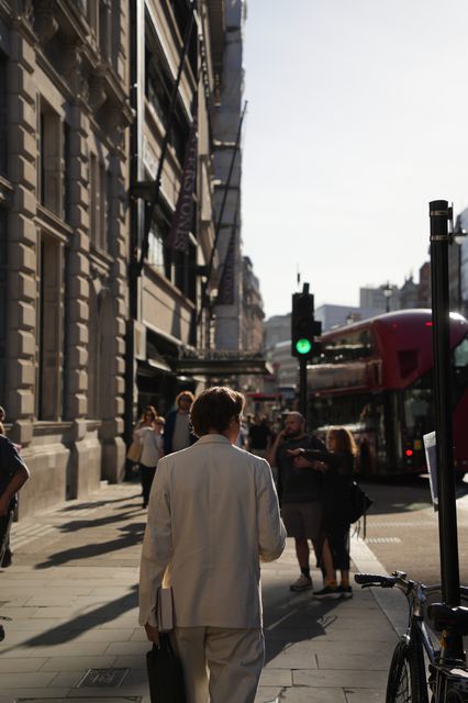A man in a pale suit walking away down a sunlit city pavement