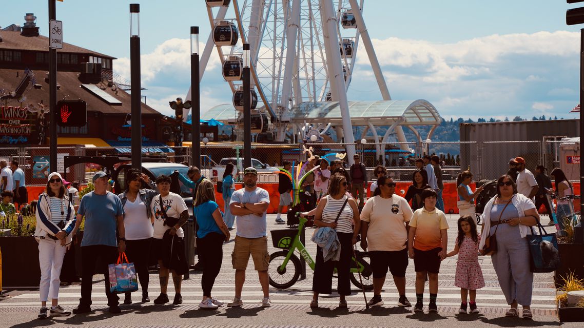 A crowd waiting at a waterfront crossing with a Ferris wheel behind
