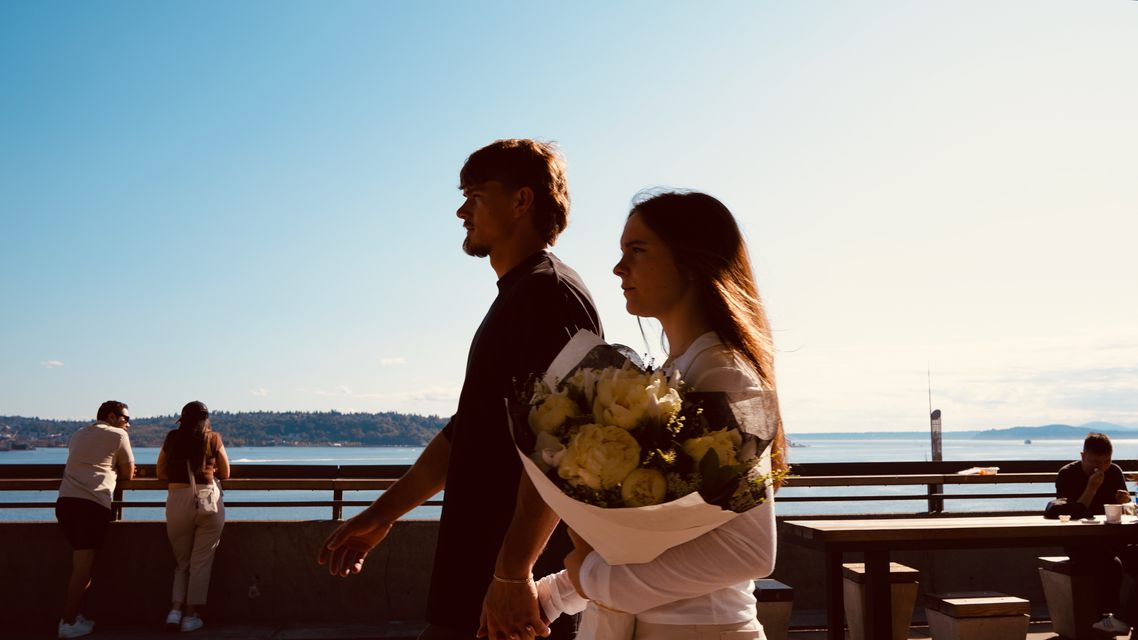 A couple walking along the waterfront at golden hour, one carrying a bouquet