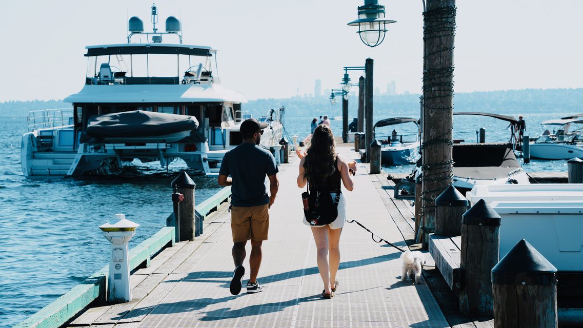 Two people and a small white dog walking down a marina dock toward a large yacht