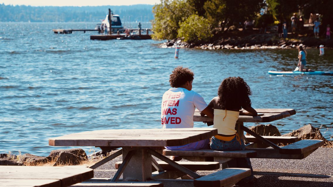Two people at a picnic table beside a sparkling lake, a paddleboarder beyond