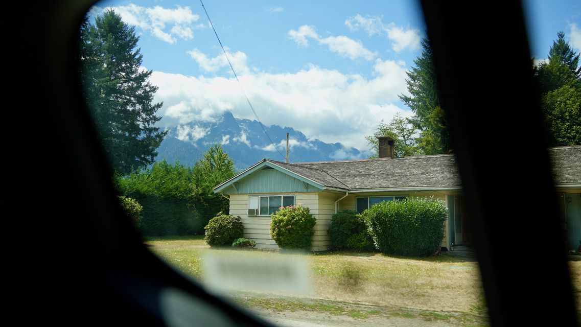 A small house with jagged mountains behind, framed by a car window