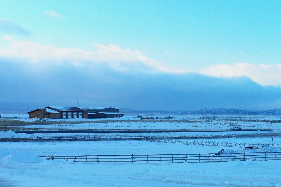 A low ranch building and fences on a snowy plain under blue clouds