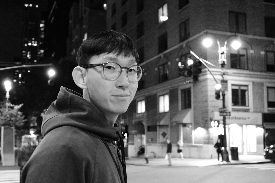 Yunzhi Li in round glasses and a hooded jacket, looking over his shoulder at the camera on a city street corner at night, in black and white.